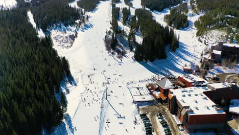 people skiing and riding chairlifts down ski slope next to parking lot and pine tree forest and snow