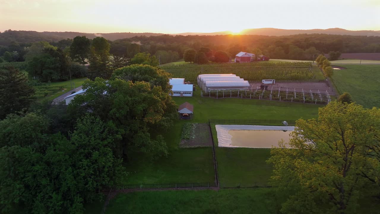 Aerial approaching shot of american farm with, stable, pond and greenhouse. GOlden sunset behind hills and forest trees in spring. Quiet rural farmstead neighborhood.