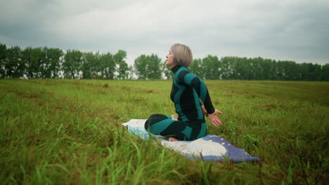 side view of old woman sitting on mat in sukhasana pose with eyes closed, practicing yoga in a vast grassy field under a cloudy sky, with trees lining the horizon, embracing tranquility