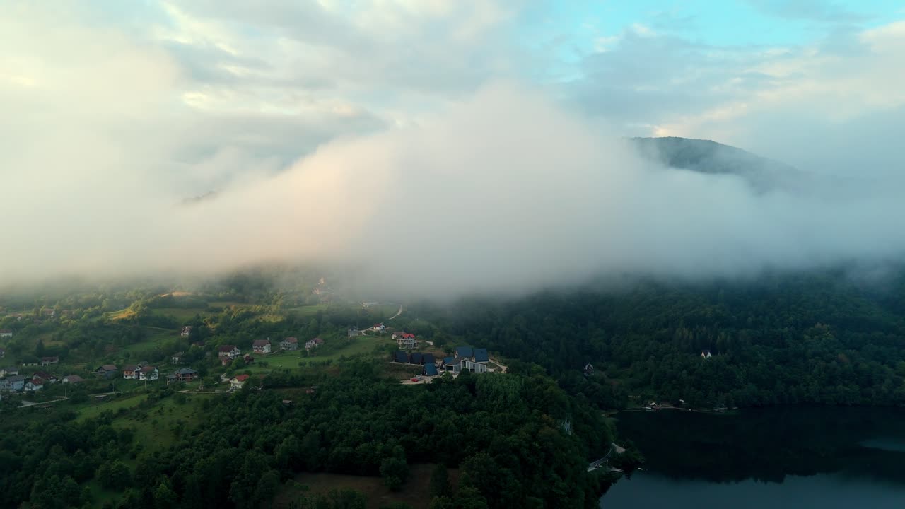 Aerial drone view of Lake Plivsko near Jajce, Bosnia and Herzegovina, covered in morning clouds and mist over forested hills, reflecting soft light and tranquil atmosphere