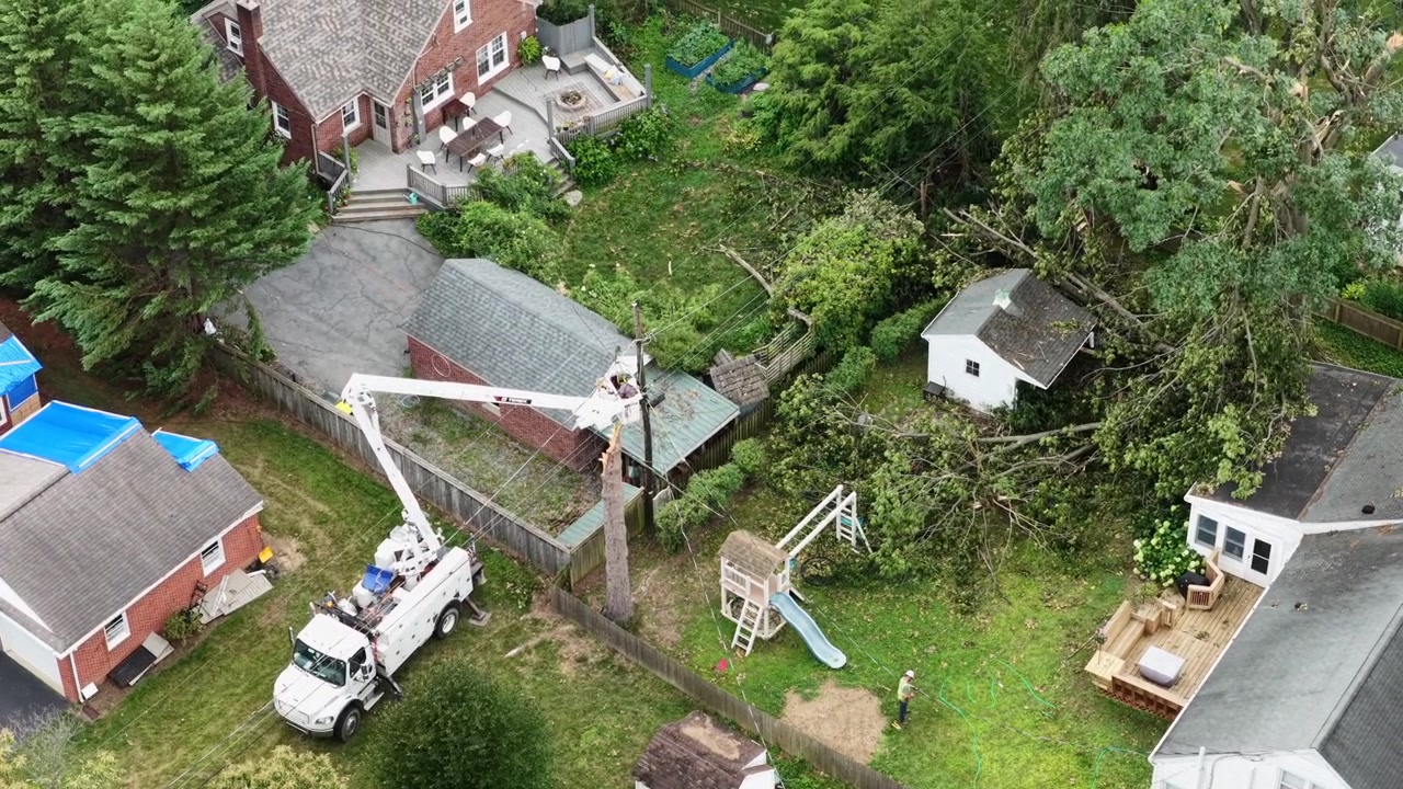 Linjearbejder i liftbil reparerer knækket elmast ved siden af et blåt presenningdækket tag i Manheim Township, Pennsylvania, med faldne trægrene spredt nær et gyngestativ i baghaven. Luftfoto fra drone, set direkte ned.