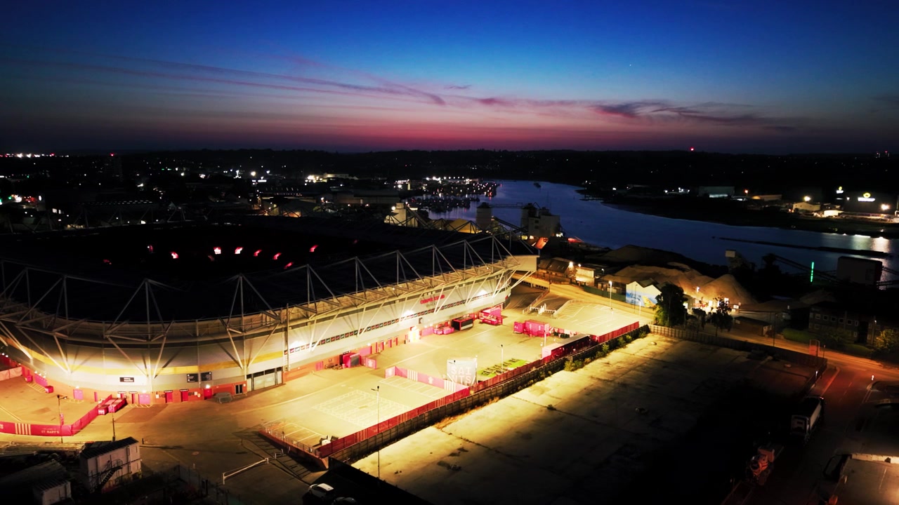 Low drone aerial of the floodlit service yard and exterior of St Marys Stadium beside the River Itchen during deep blue hour
