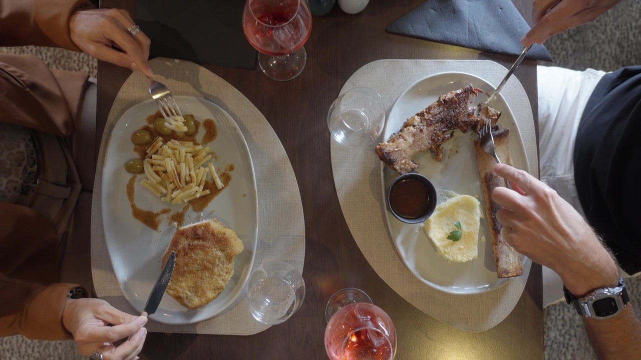 Two people eat lunch at a restaurant table with meat, sides, and wine glasses