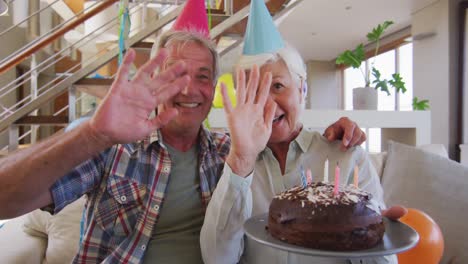 Portrait-of-senior-caucasian-couple-holding-cake-celebrating-birthday-waving-looking-at-camera-at-ho