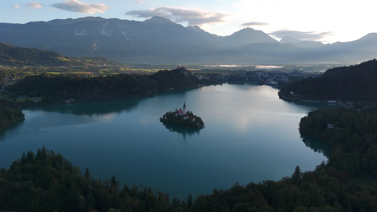 Ampia vista aerea del Lago di Bled al crepuscolo, Slovenia, con la chiesa sull'isola centrata in acque calme, nuvole soffici sopra le montagne alpine e una tenue luce serale. Atmosfera tranquilla in una giornata di sole.