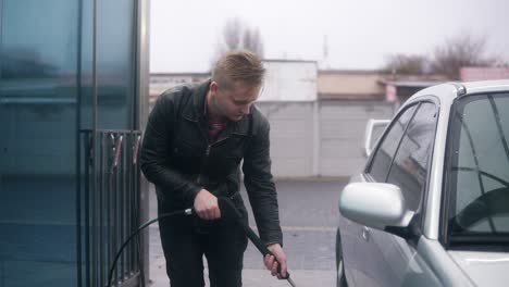 Young-attractive-stylish-man-washing-his-silver-sportcar-with-water-jet-on-self-service-carwash.-He-is-carefully-cleaning-the-tyres-of-the-car.-Slowmotion-shot