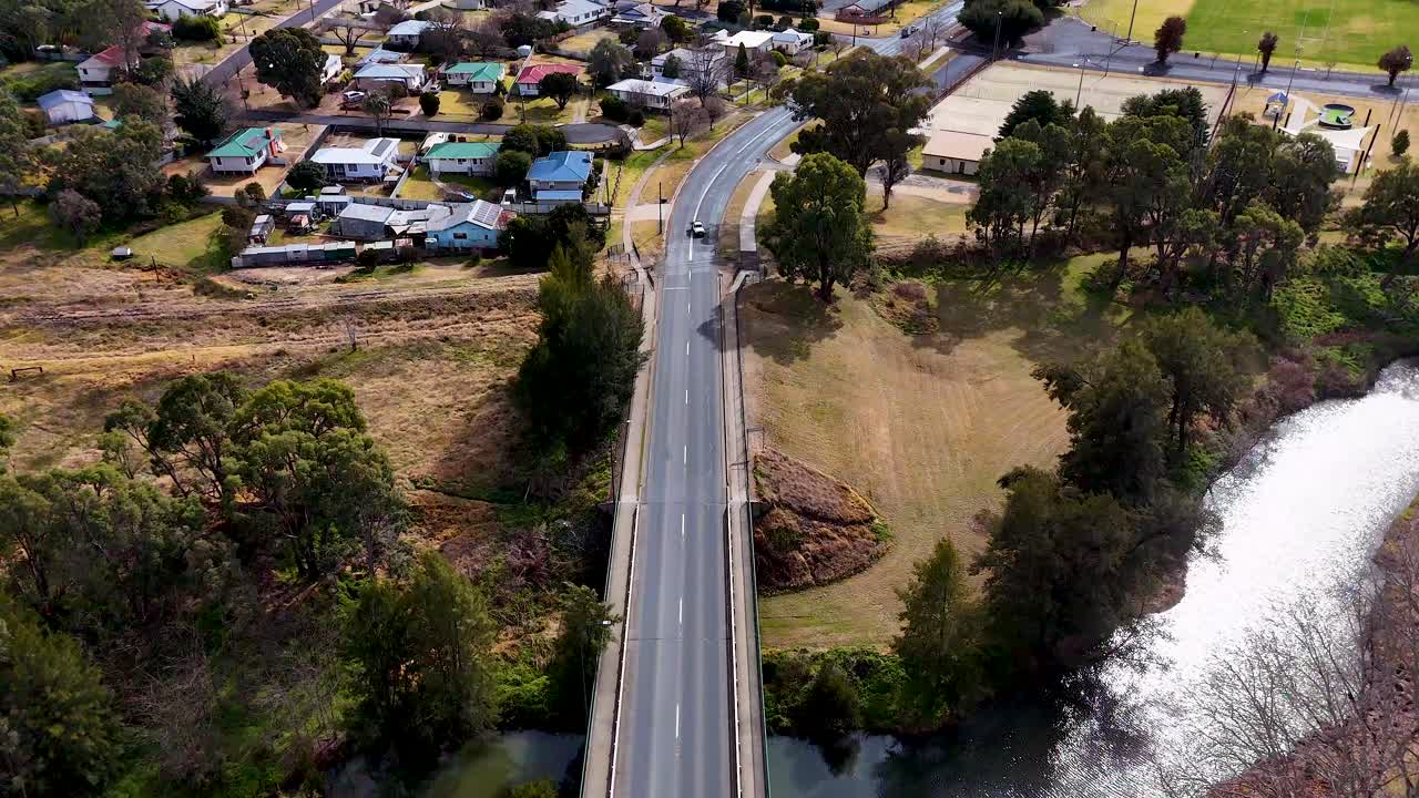 A single car travels across a road bridge spanning a riverbank, surrounded by trees and residential homes, captured in daylight from a high aerial perspective