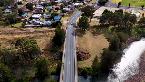 a single car travels across a road bridge spanning a riverbank, surrounded by trees and residential homes, captured in daylight from a high aerial perspective