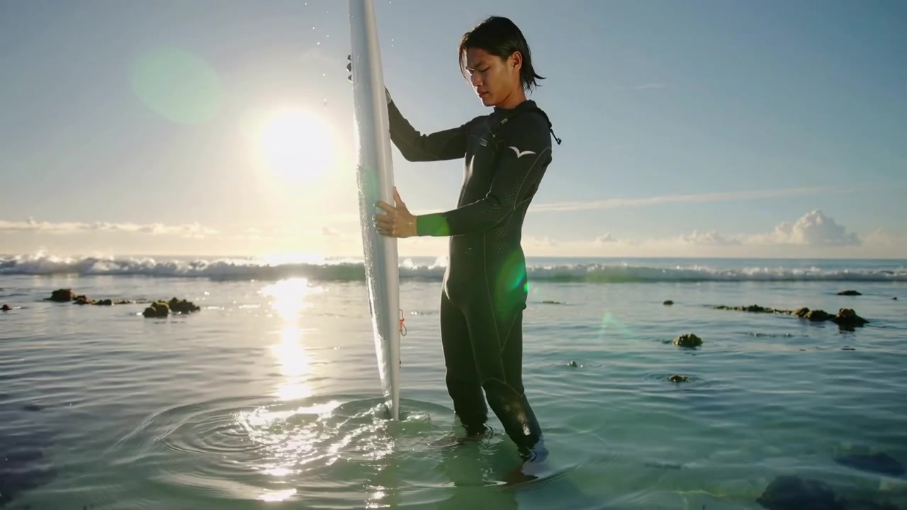 Surffaaja mustassa märkäpuvussa tarkkailee aaltoja, säätää otettaan ja nostaa valkoista surffilautaa matalassa vedessä.