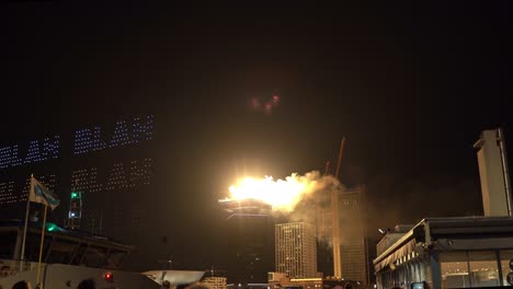 Espectáculo-De-Drones-Brillantes-Con-Letras-Escritas-En-El-Cielo-Y-Fuegos-Artificiales-Sobre-El-Horizonte-Urbano-De-La-Ciudad-Por-La-Noche