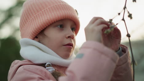 side close-up of a little girl dressed in a pink jacket and beanie, intently examining and touching a dry tree branch