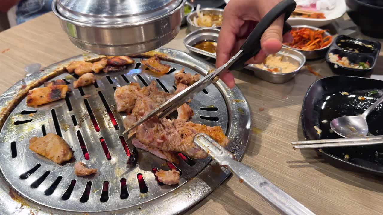 Hands skillfully cut grilled pork on a Korean BBQ grill in a lively Bangkok setting, highlighting culinary precision and vibrant dining