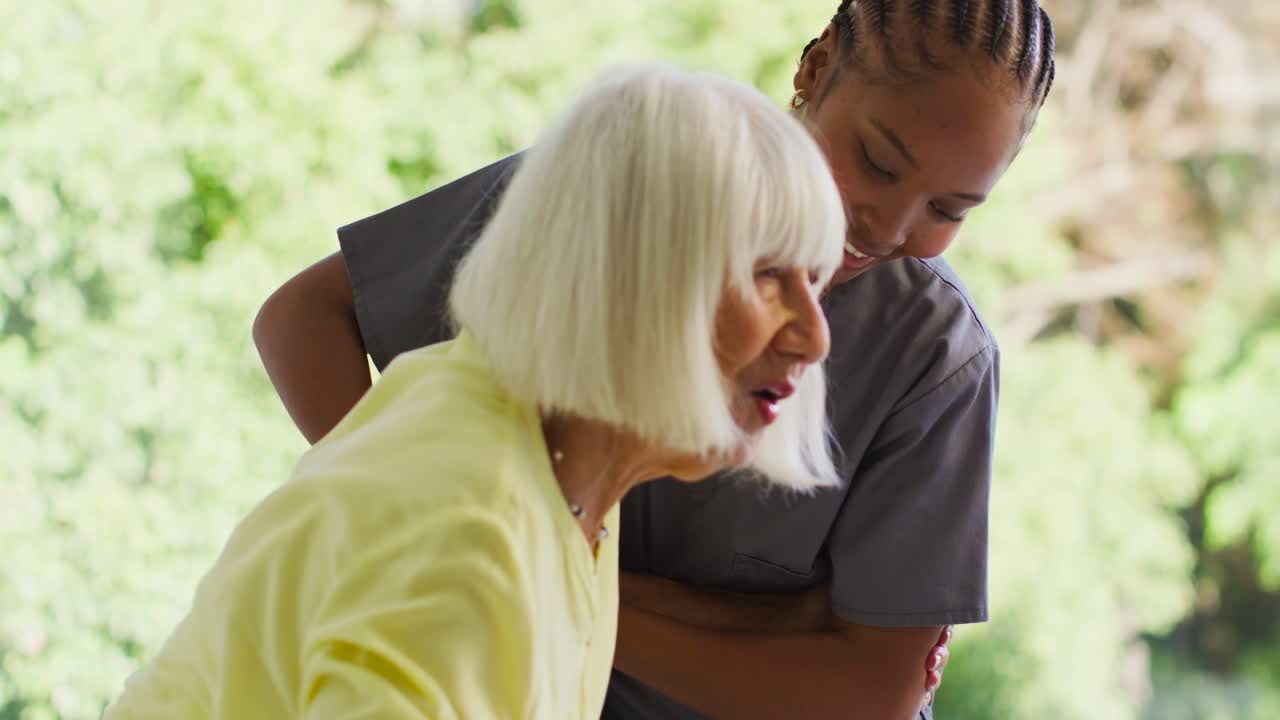 Assistente afroamericana in divisa medica che aiuta una donna anziana ad alzarsi con il bastone, stabilizzandola vicino alla finestra