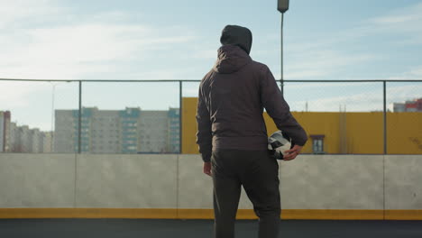 back view of person holding soccer ball while entering sports arena, showcasing urban environment with soft natural lighting, the individual is dressed in casual athletic wear