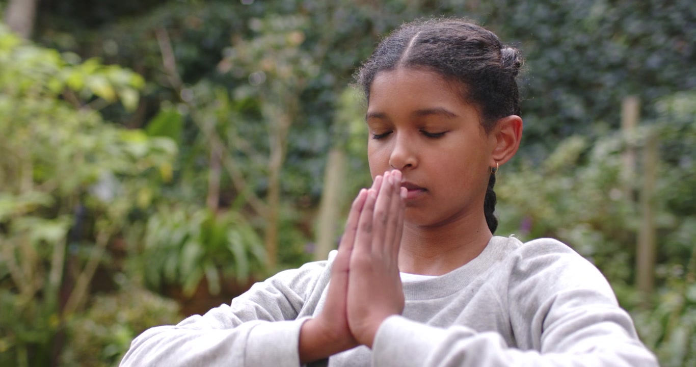 African American teen standing in garden starting raising arms, pressing palms to chest, meditating