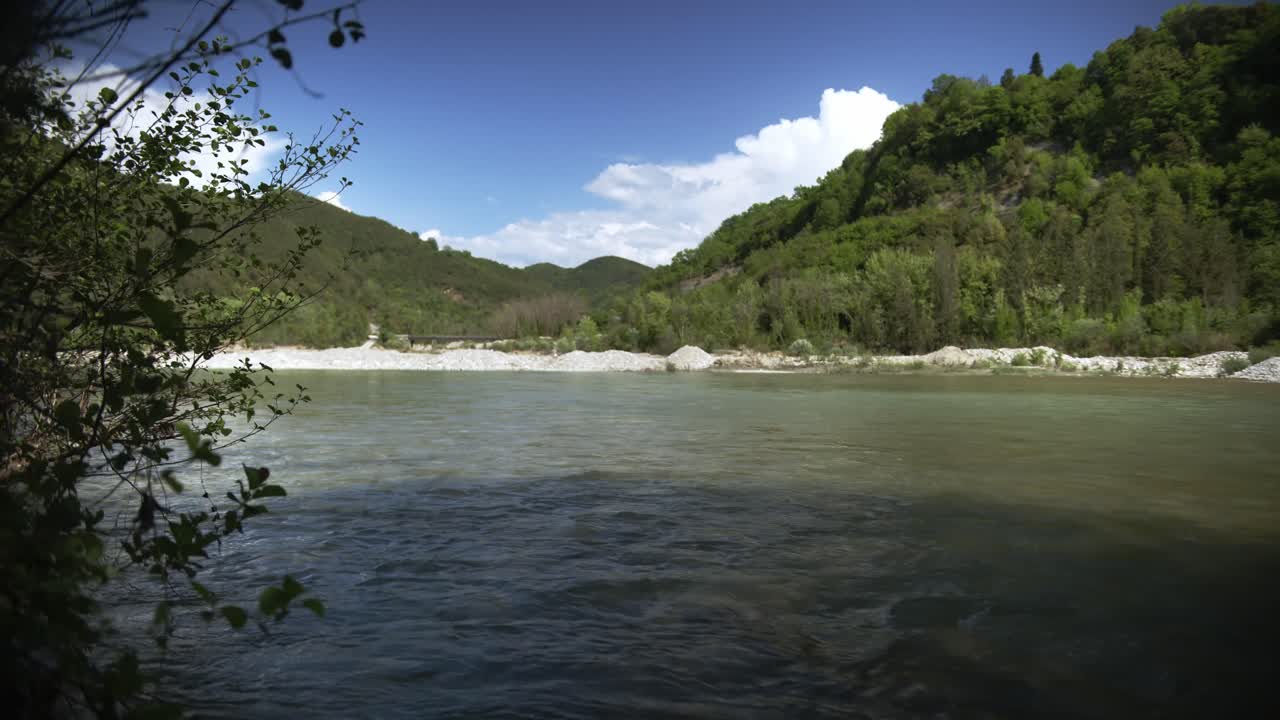 Tranquil wide-angle shot of a river with forested hills and cloudy blue sky in the background. Captured in Tzoumerka. Perfect for nature documentaries, travel scenes, or background visuals.