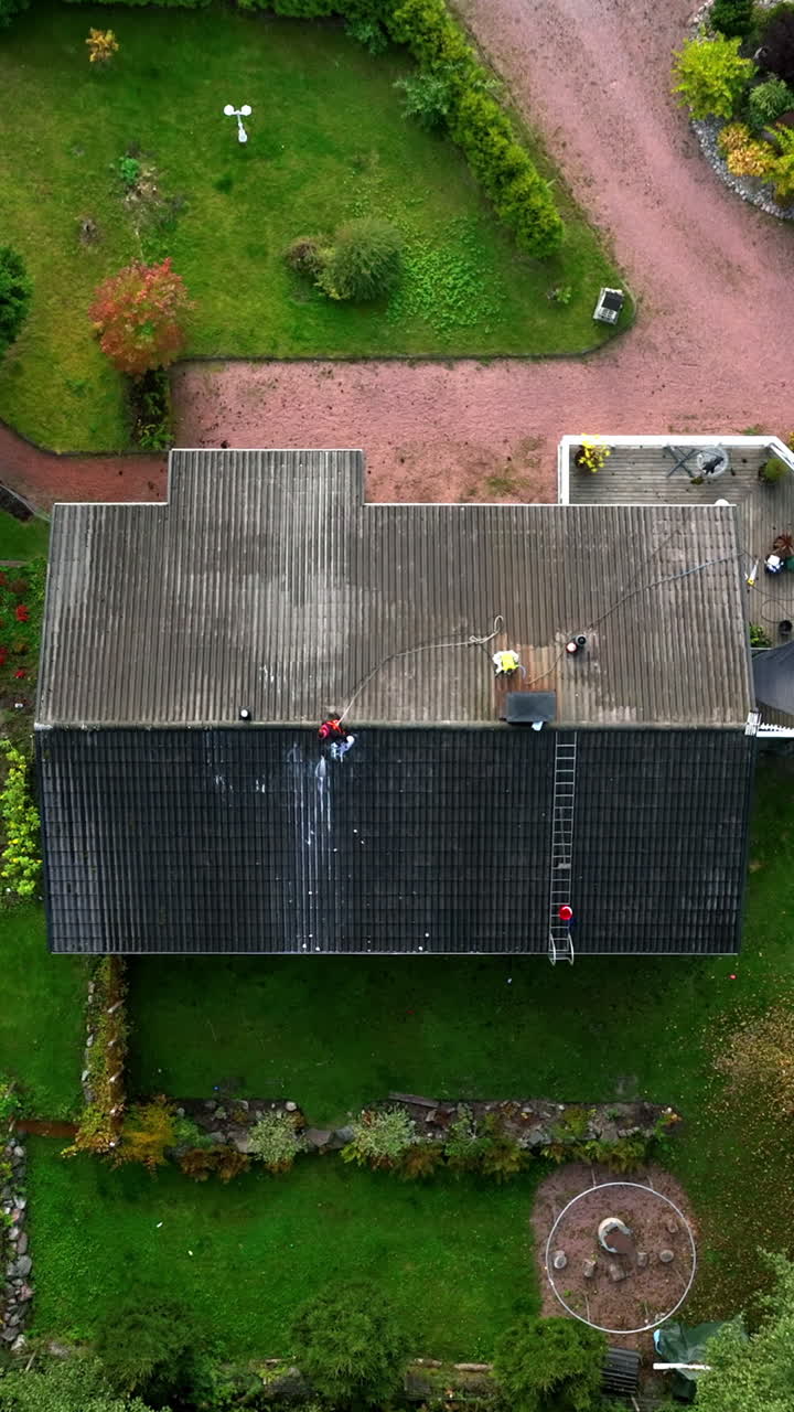 Vertical drone shot above a woman pressure washing tiles of a house roof