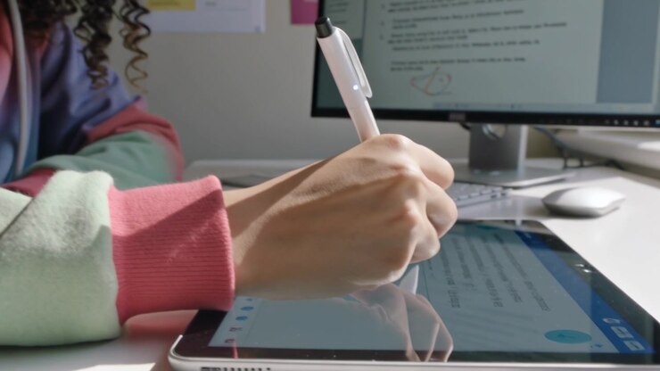 Picking up student wearing pastel hoodie placing stylus on tablet at desk, writing from monitor