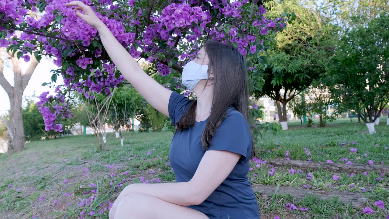 mujer caucásica con mascarilla aislada en el parque con el pelo moviéndose en el viento recogiendo flores rosas del árbol
