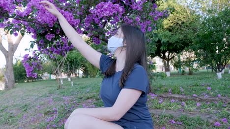 Caucasian woman with face mask isolated in the park with hair moving in the wind collecting pink flower from the tree