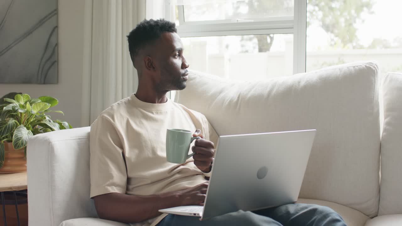 African man sitting on sofa, holding mug, typing on laptop, resting eyes while gazing out window