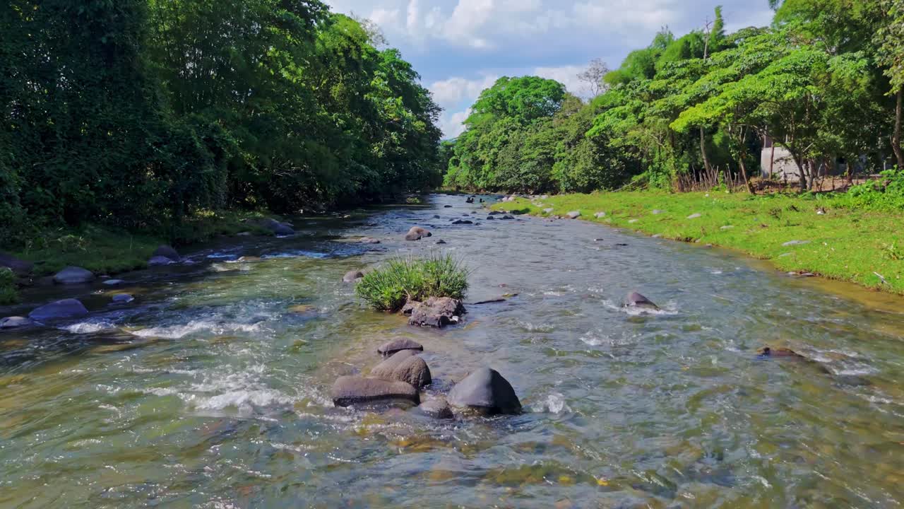 Smooth aerial drone footage flying forward low above the flowing water of Rio Maimon, surrounded by lush green trees and riverbanks on sunny day. Nature, travel, serene river scene