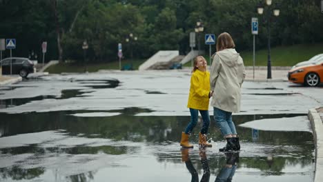 happy blonde woman with her teenage daughter in a yellow jacket jumping in a puddle holding hands in the park after the rain