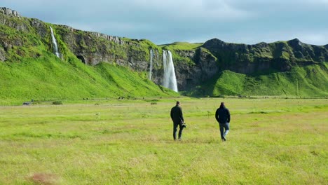 two tourists walking towards seljalandsfoss and gljufrabui waterfalls over a beautiful field of grass on a sunny day in southern iceland