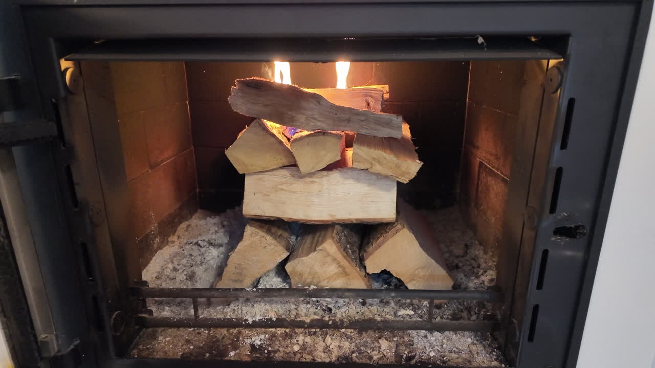 A stacked arrangement of firewood burns inside a closed indoor fireplace, with small flames visible at the top of the wooden logs. Ash and embers cover the base of the firebox