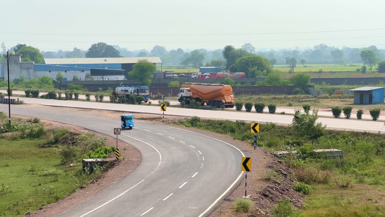Static shot of light traffic moving along a rural highway, tankers and vehicles passing through open farmland under soft daylight with distant trees and structures