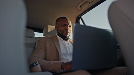 a confident brunette man with black skin in a brown suit works on a gray laptop during his business trip outside the urban area