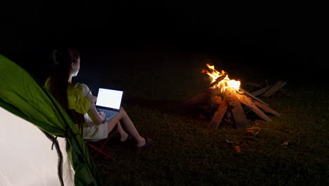 Junge-Frau-Bleibt-In-Abgelegenen-Gegenden-In-Verbindung,-Während-Sie-Nachts-Im-Freien-Campt-Und-Vor-Dem-Lagerfeuer-Am-Laptop-Arbeitet