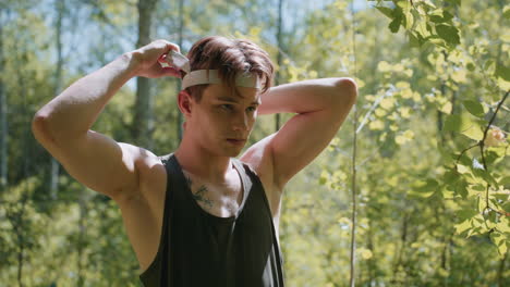 front view of tourist vigilantly observing environment with chest tattoo and bare shoulder visible, brown hair falling over brow, dappled sunlight filtering through green leaves in forest clearing