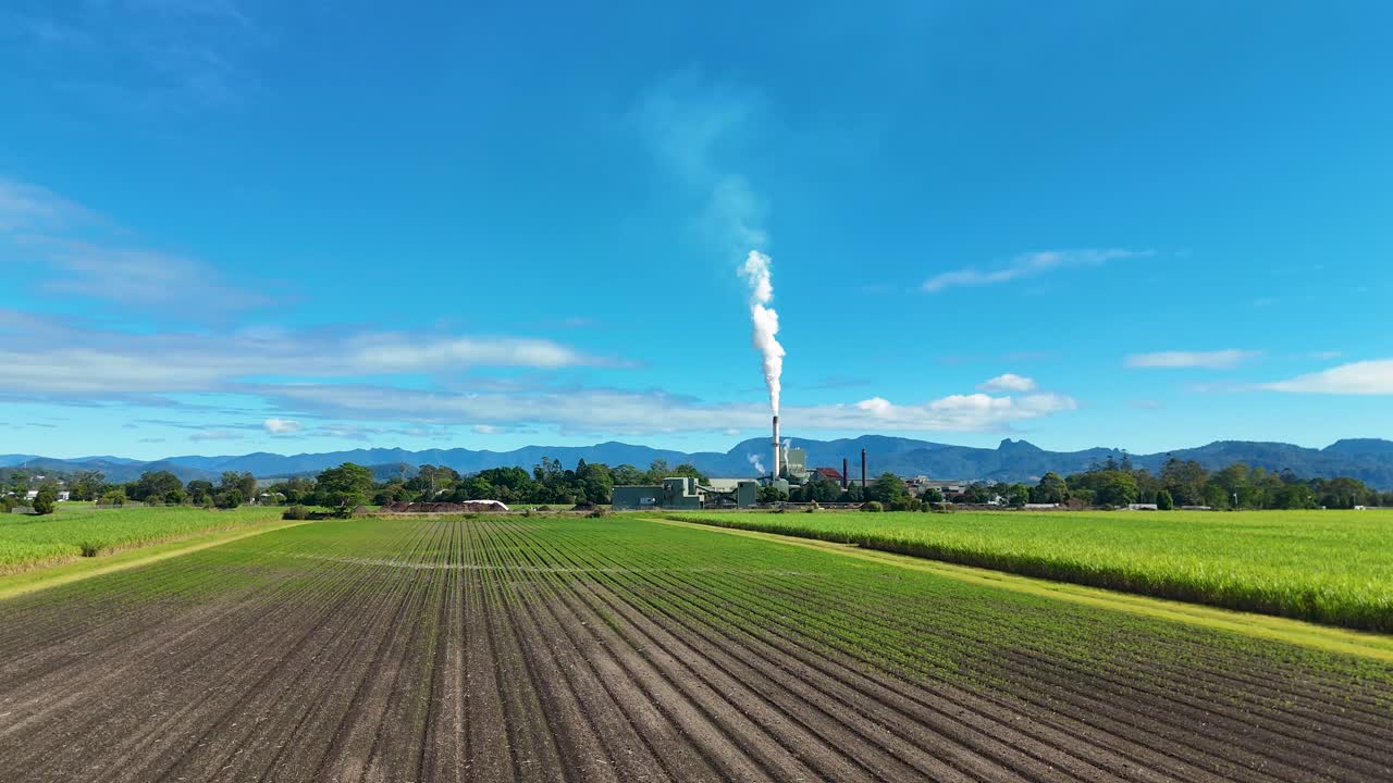 Smoke billows from a tower over expansive farmland, with clear blue skies and distant mountains in the background