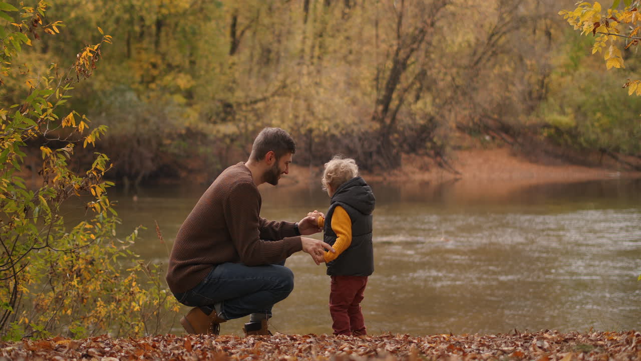 padre feliz está hablando con su hijo hijo en el bosque de pie en la costa de un lago o río en el fin de semana de otoño viaje familiar en el bosque o parque de reserva