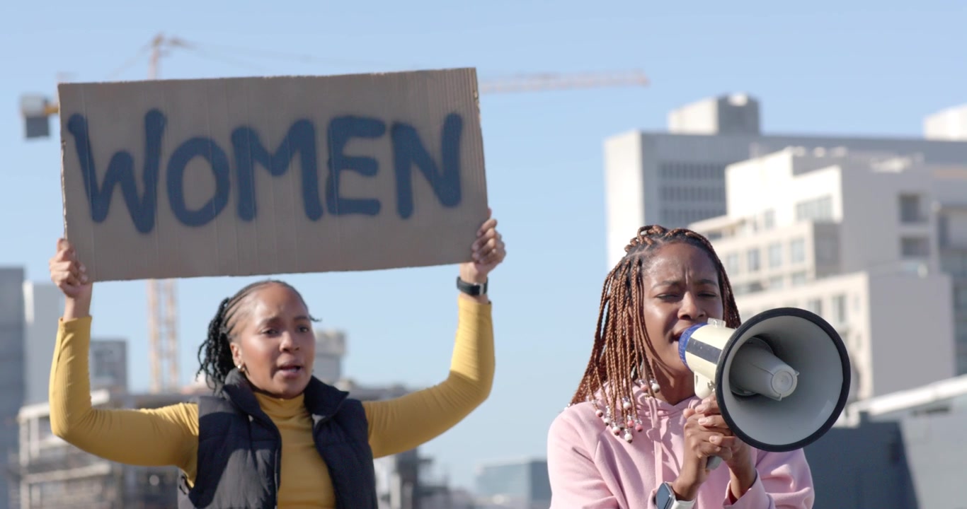 Afrikanische Frauen protestieren auf dem Dach, die rechte Frau hebt ein Megafon und ruft, hält ein „WOMEN“-Schild und trägt einen Kapuzenpullover.