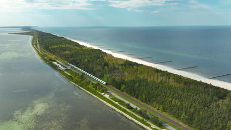 A-spectacular-aerial-view-of-Kuźnica-on-the-Hel-Peninsula,-a-train-moving-alongside-the-narrow-strip-of-land-flanked-by-the-bay-and-the-Baltic-Sea,-highlighting-the-area's-unique-geography