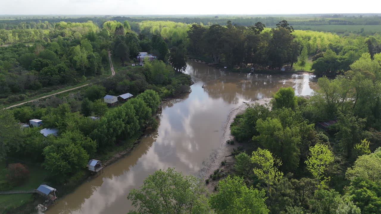 Drone footage showcases a serene rural school and charming cabins nestled along a winding river in the lush Argentine Delta landscape. A peaceful depiction of remote riverside living and nature.
