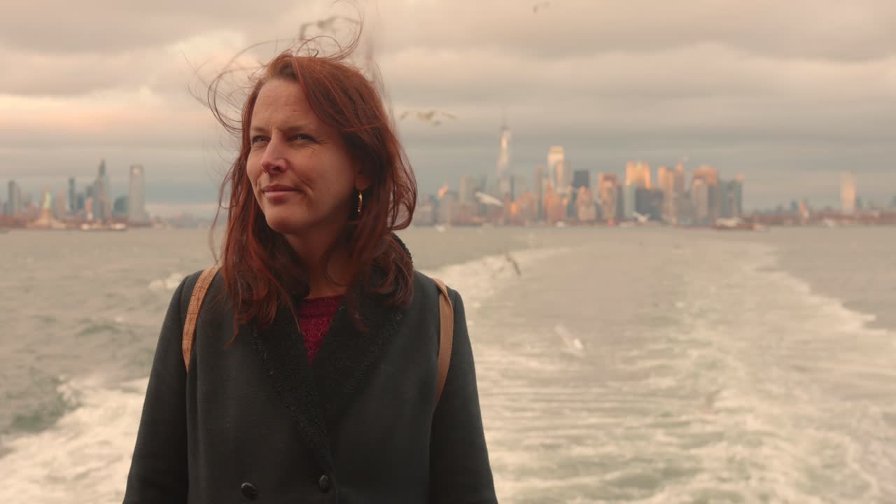 A contemplative woman with wind-blown hair looks towards the beautiful Manhattan skyline and Statue of Liberty from the back of a ferry. Captures themes of freedom, travel, and urban exploration