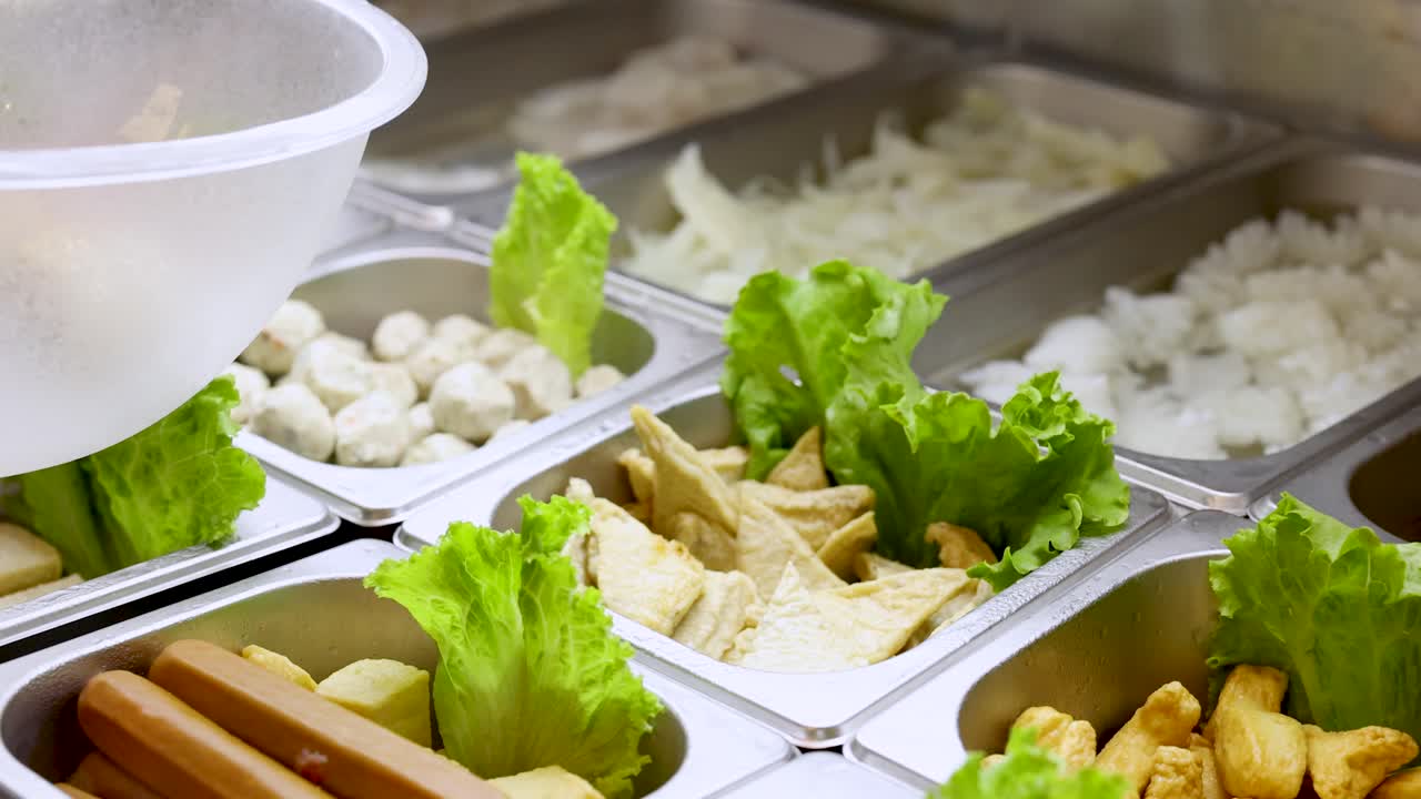 Hand selects noodles from buffet with tofu, fish balls, vegetables under bright indoor lighting