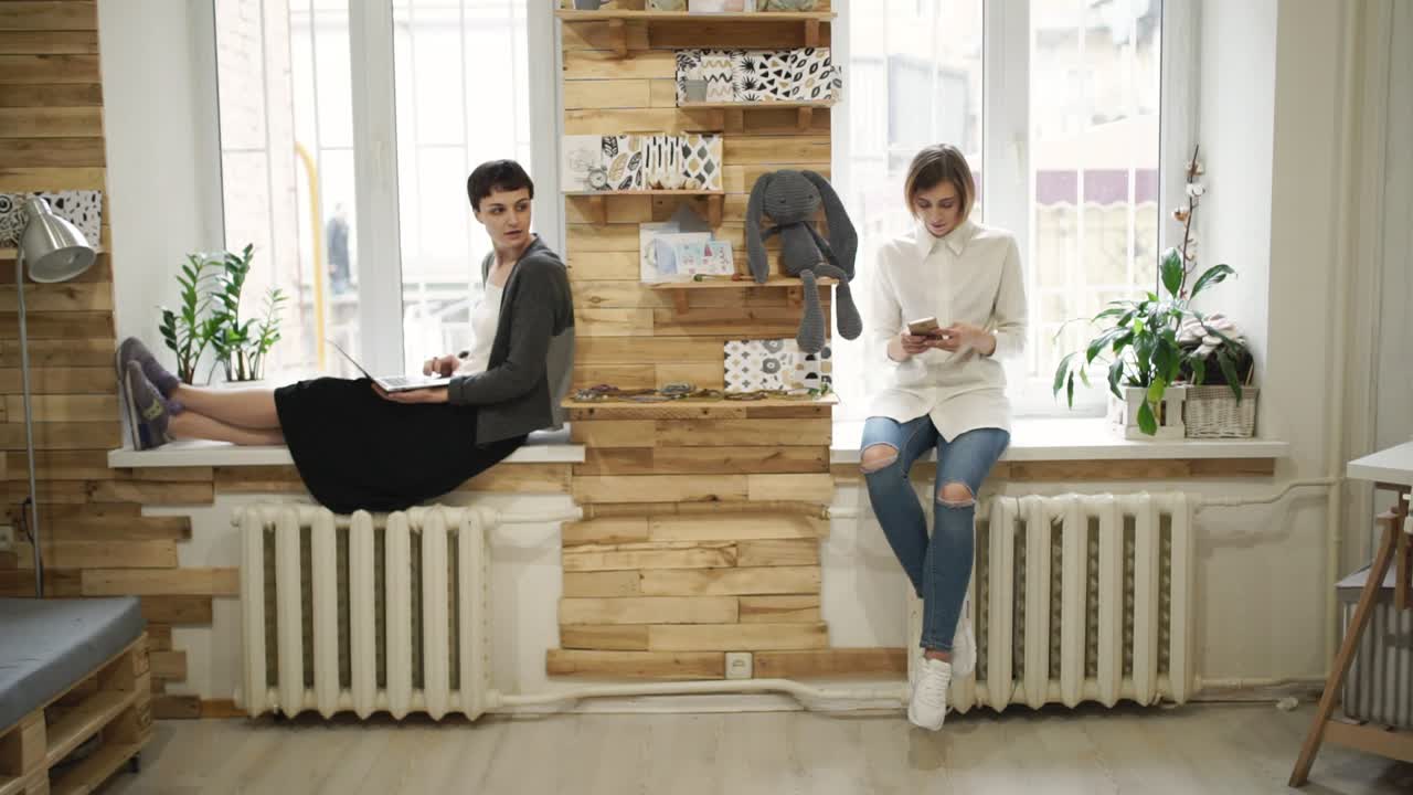 유리창 위에 앉아 있는 노트북을 사용하는 젊은 여성. 휴대폰을 사용하는 여성