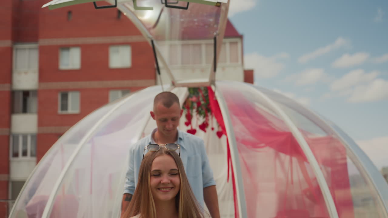 coppia sorridente in una cupola a bolla panoramica sotto un drappeggio rosso e un baldacchino floreale, donna con trecce e top bianco, partner che sbircia da dietro, skyline urbano sullo sfondo, luce solare dorata, momenti di gioia spontanei