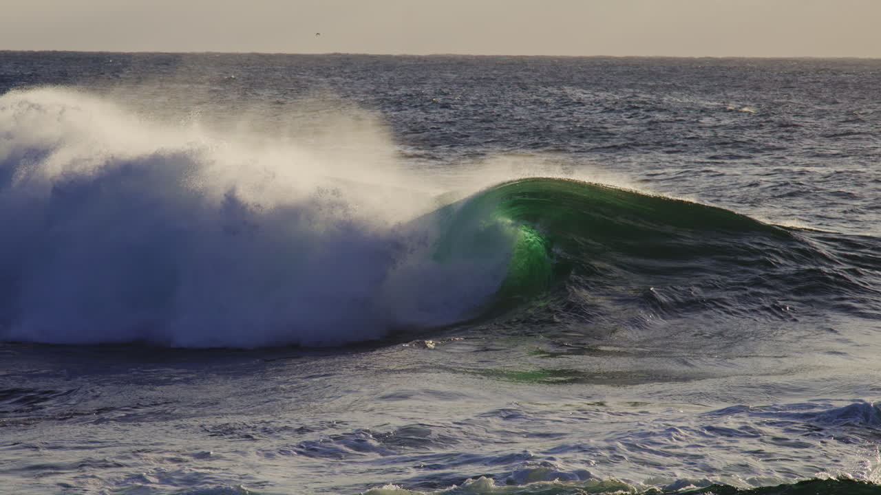Backlit green ocean wave crashes and barrels with white foam ball and mist rising off top at golden hour sunset