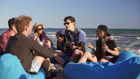 young man playing guitar among group of friends sitting on easychairs on the beach and singing on a summer evening. slowmotion shot.