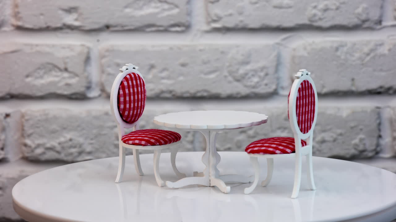 Carved wooden toy table and two chairs at both sides rotating. Pretty furniture for a dolly house. White table and red-and-white chairs against brick wall backdrop.