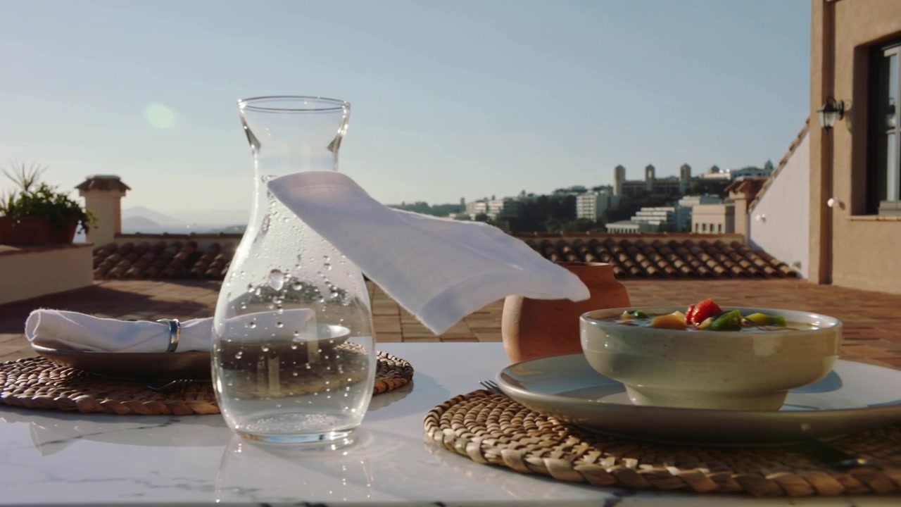 Una leggera raffica solleva un tovagliolo portandolo attraverso una caraffa su un tetto, mentre un avambraccio posiziona una ciotola per servire.