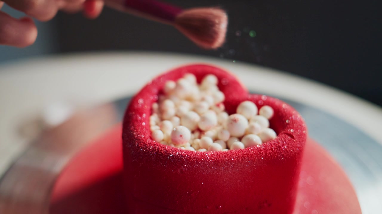 Una mano usa un pennello per spolverare zucchero su una torta rossa a forma di cuore. Decorazioni bianche a forma di perla riempiono il centro del cuore. Il processo è ravvicinato, lento e preciso. Ideale per San Valentino.