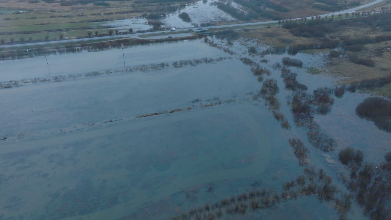Aerial establishing view of high water, Durbe river flood, brown and muddy water, agricultural fields under the water, overcast winter day with light snow, drone shot moving forward tilt down