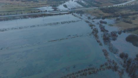 Luftaufnahme-Von-Hochwasser,-Überschwemmung-Des-Durbe-Flusses,-Braunem-Und-Schlammigem-Wasser,-Landwirtschaftlichen-Feldern-Unter-Wasser,-Bewölktem-Wintertag-Mit-Leichtem-Schneefall,-Drohnenaufnahme,-Die-Sich-Nach-Vorne-Bewegt-Und-Nach-Unten-Neigt