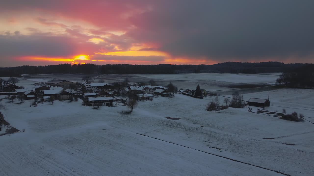 un pueblo rural rodeado de árboles en invierno en baviera, alemania, con nieve en los campos y techos vistos desde arriba, imágenes aéreas de drones mientras se pone el sol con nubes rojas y cielo